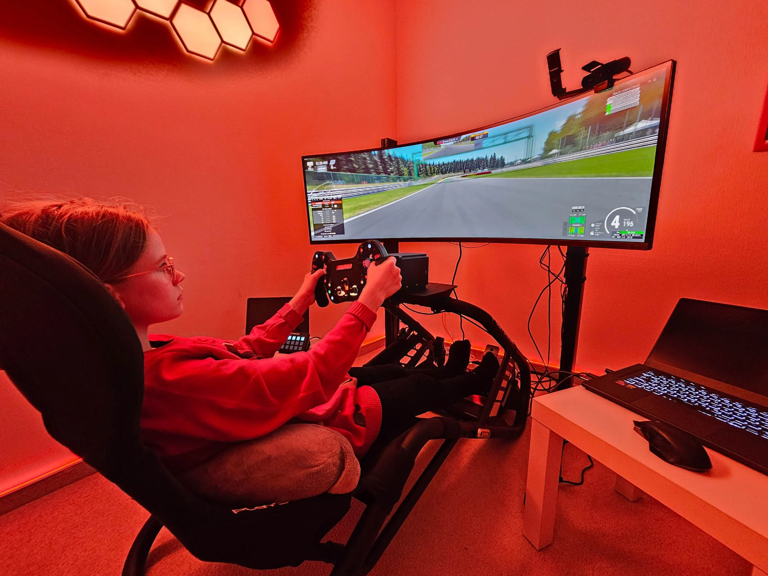 simracingpeterlauritisgt3simulation003 | Lauritisde Person in a Red Hoodie Uses a Steering Wheel Controller to Play a Racing Game on a Large Monitor in a Red lit Room