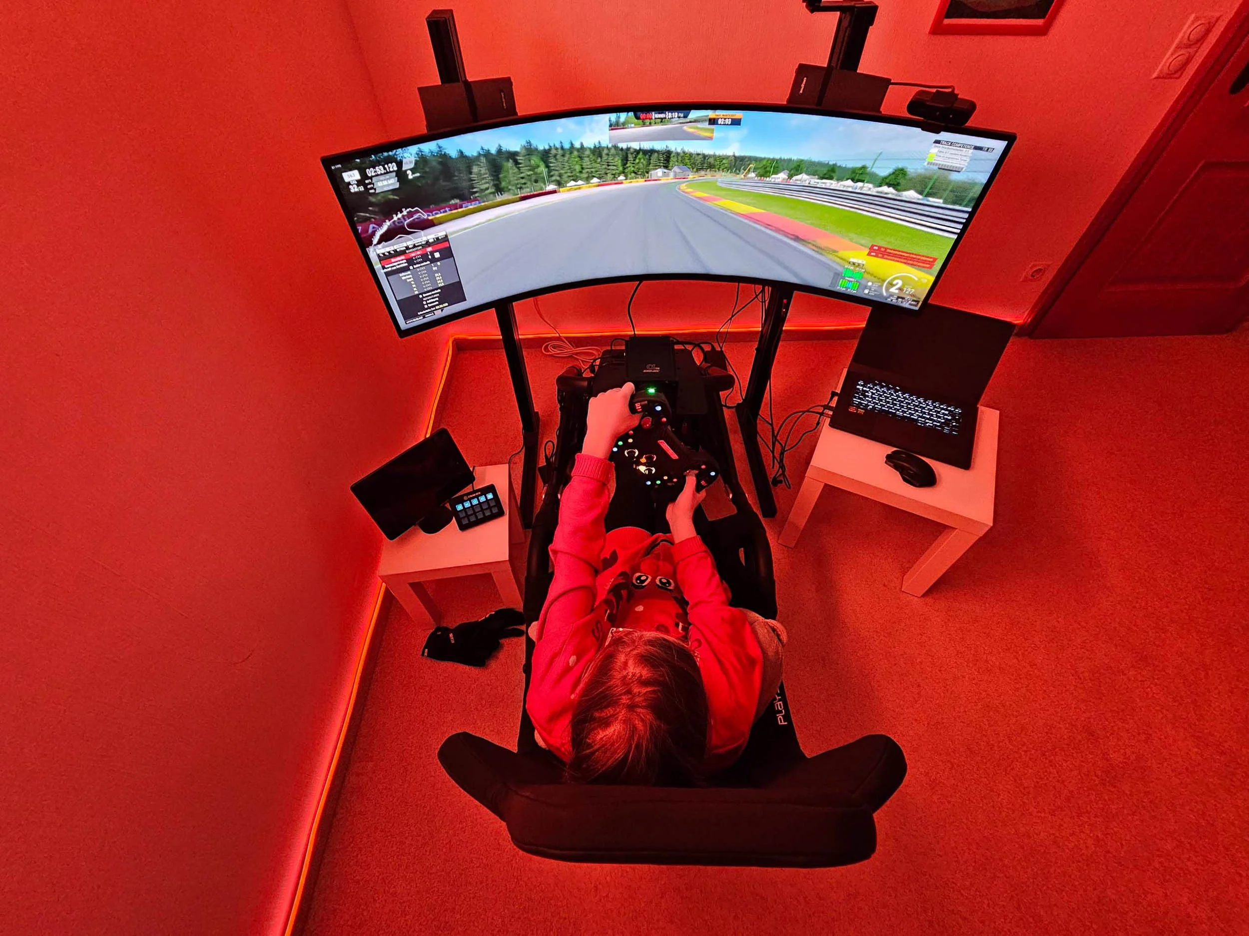simracingpeterlauritisgt3simulation004 | Lauritisde Person Seated at a Curved Multi monitor Racing Simulator in a Red lit Room Hands on a Steering Wheel with Pedals Beneath Laptop on a Nearby Table Nearby