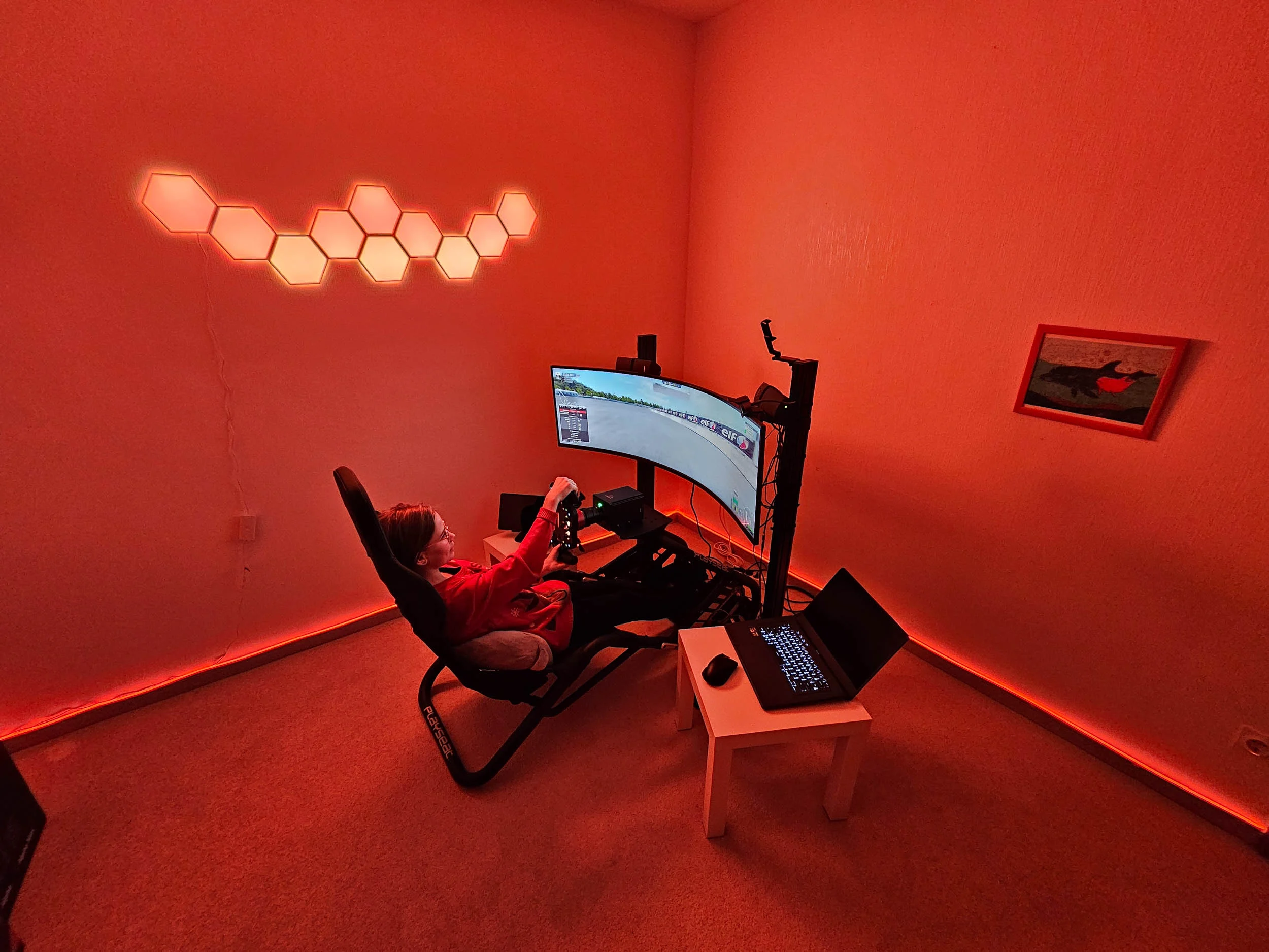 simracingpeterlauritisgt3simulation005 | Lauritisde Person in a Red lit Room Using a Home Racing Simulator with a Curved Monitor Wheel Pedals and a Laptop on a Small White Table