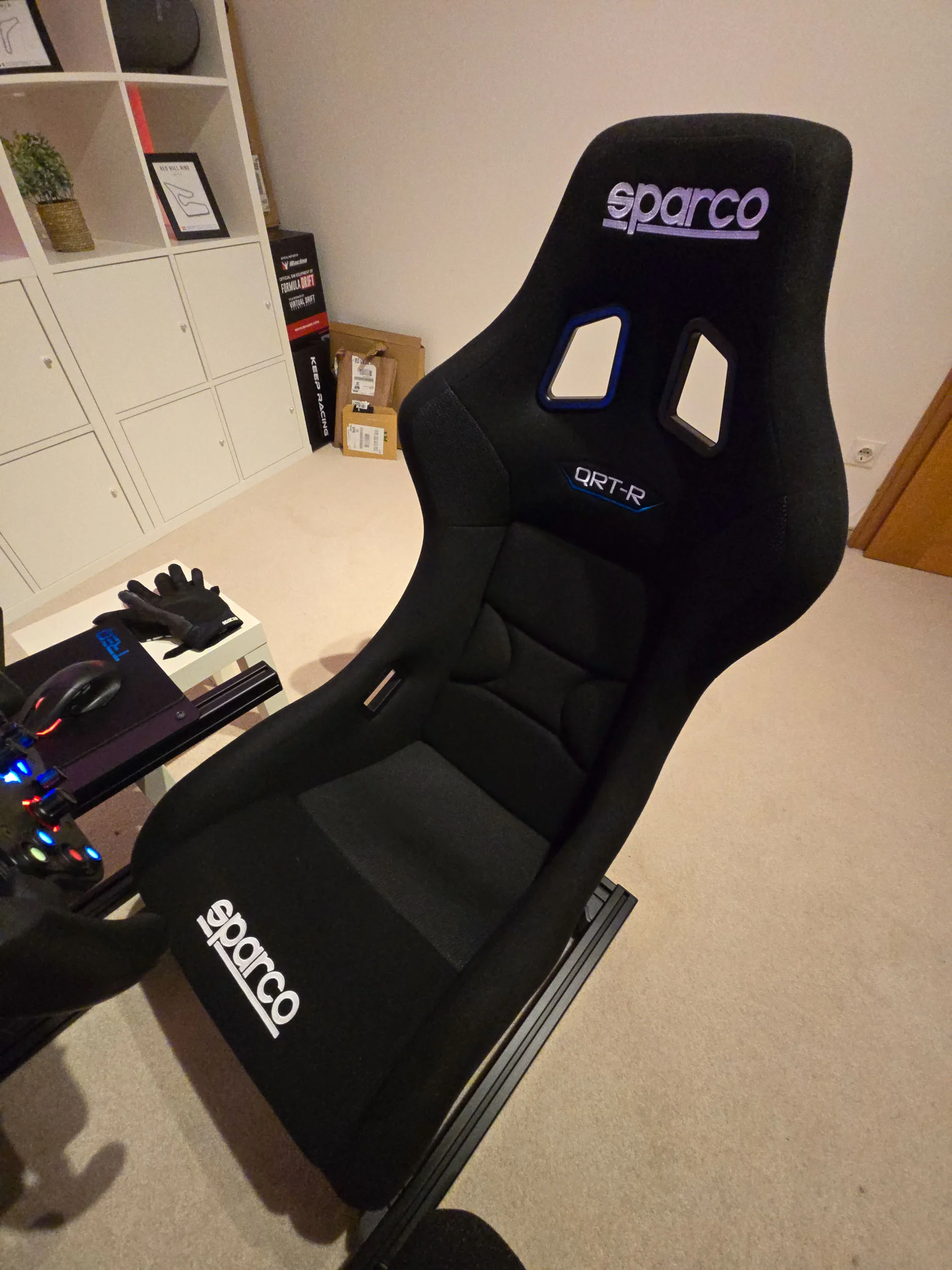 simracingpeterlauritisgt3simulation013 | Lauritisde Black Sparco Race Seat with Blue Harness Holes in a Home Office White Shelves and a Desk with Gloves Nearby
