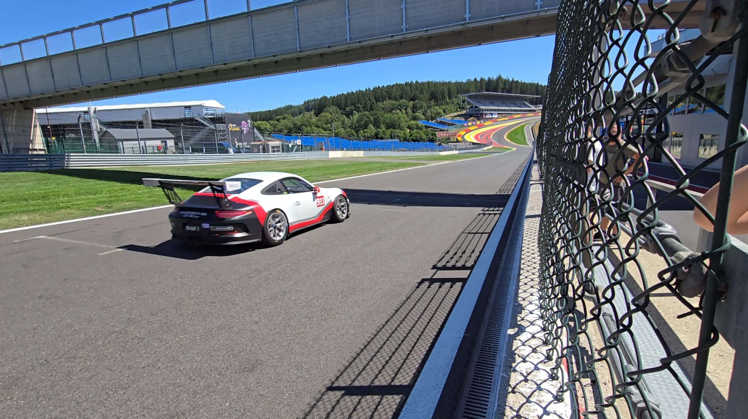 Race Car on a Dark Asphalt Track Near Barriers with Colorful Curbs in the Distance and a Chain Link Fence in the Foreground Under a Bridge